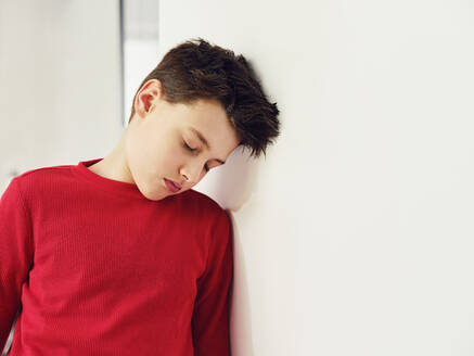 Tired boy with eyes closed leaning head on wall at home - PWF00354