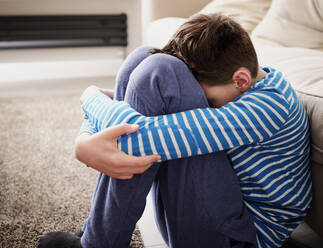 Sad boy hugging knees at sofa in living room - PWF00356