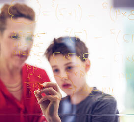 Boy writing on glass by mother while learning mathematics at home - PWF00372