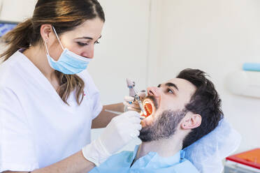 Female dentist wearing protective face mask examining male patient's teeth at medical clinic - WPEF04469