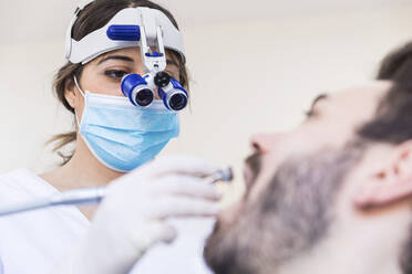 Zahnärztin mit Lupenbrille bei der Arbeit an den Zähnen eines männlichen Patienten in einer medizinischen Klinik - WPEF04478