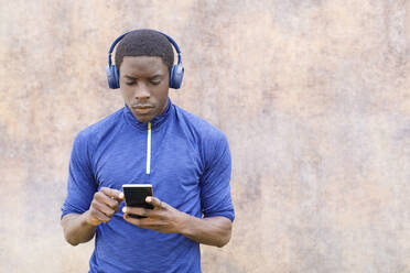 Man using mobile phone while listening music through headphones in front of wall - IFRF00626