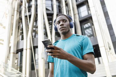 Man wearing wireless headphones holding mobile phone in front of building - IFRF00637