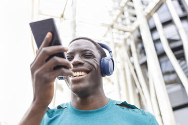Lächelnder junger Mann mit Kopfhörern und Smartphone vor einem Gebäude - IFRF00640