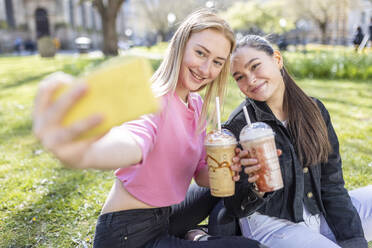 Lächelnde Freundinnen, die ein Selfie mit ihrem Handy machen, während sie im Park einen Milchshake trinken - WPEF04512