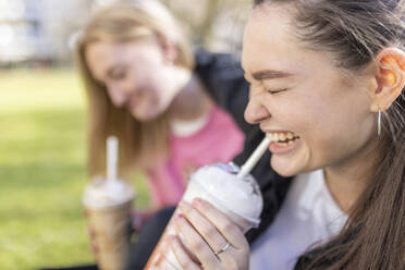 Happy woman drinking milkshake with female friend in background at public park - WPEF04514