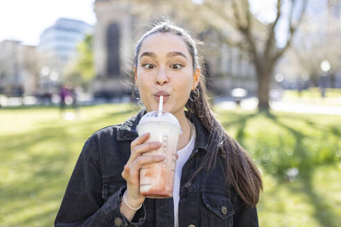 Junge Frau mit schielendem Blick beim Trinken eines Milchshakes im Park - WPEF04516