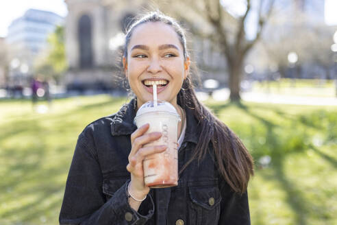 Smiling woman looking away while drinking milkshake at public park - WPEF04517
