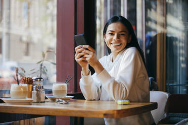 Glückliche Frau sitzt mit Smartphone in einem Café - OYF00354