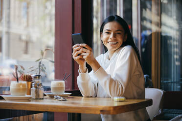 Happy woman sitting with smart phone at coffee shop - OYF00354