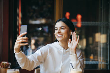 Cheerful woman waving on video call while sitting in cafe - OYF00357