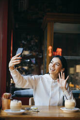 Smiling woman waving on video call at coffee shop - OYF00358