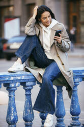 Lächelnde Frau, die ein Mobiltelefon benutzt, während sie auf einem blauen Geländer sitzt - OYF00365