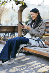 Lächelnde Frau im Mantel, die ihr Smartphone benutzt, während sie auf einer Bank im öffentlichen Park sitzt - OYF00369