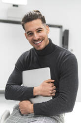 Lächelnder junger Geschäftsmann mit Laptop im Büro - JAQF00647