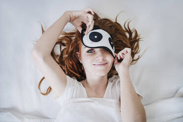 Lächelnde Frau nimmt die Augenmaske ab, während sie zu Hause auf dem Bett liegt - EBBF03546