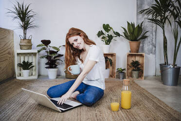 Junge Frau, die einen Laptop benutzt, während sie zu Hause auf dem Boden Kaffee trinkt - EBBF03557