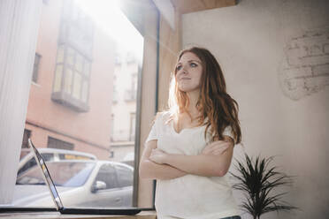 Young woman with arms crossed standing by laptop at home - EBBF03565