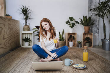 Lächelnde junge Frau sitzt im Schneidersitz und trinkt Saft vor einem Laptop auf dem Boden zu Hause - EBBF03566