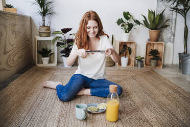 Smiling young woman photographing food while sitting on floor at home - EBBF03571