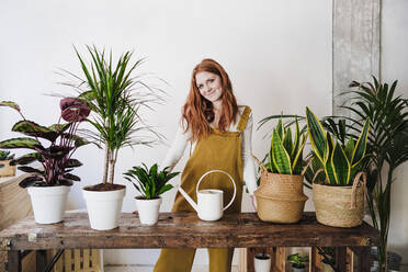 Young woman standing in front of watering can at home - EBBF03575