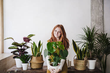 Smiling redhead woman holding potted plant at home - EBBF03579