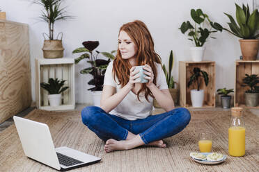 Frau mit gekreuzten Beinen, die auf einen Laptop schaut, während sie einen Kaffee auf dem Boden zu Hause trinkt - EBBF03585
