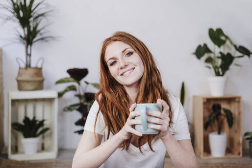 Lächelnde junge Frau hält Kaffeetasse zu Hause - EBBF03586