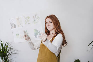Smiling redhead woman touching glass device screen in front of wall at home - EBBF03603