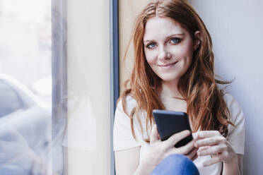 Lächelnde Frau mit Handy in der Hand, die zu Hause am Fenster sitzt - EBBF03623