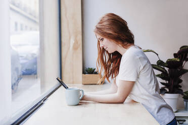 Redhead woman using mobile phone while leaning on window sill at home - EBBF03624