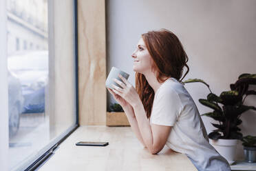 Smiling woman having coffee while leaning on window sill at home - EBBF03625