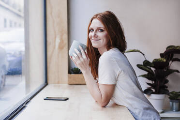 Beautiful woman having coffee leaning on window sill at home - EBBF03626