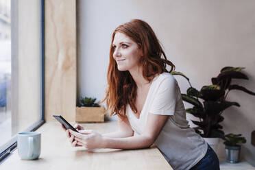 Beautiful young woman holding smart phone while leaning on window sill at home - EBBF03627