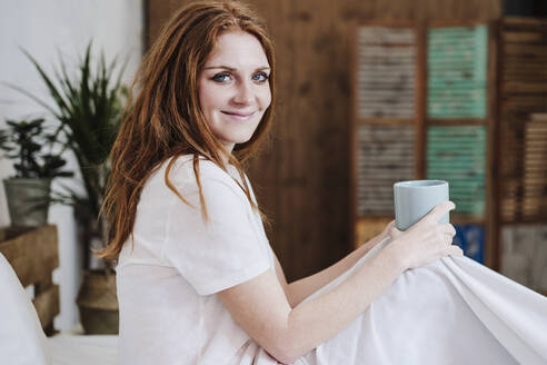 Lächelnde junge Frau mit Kaffeetasse auf dem Bett - EBBF03632