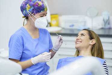 Zahnärztin mit Gesichtsschutzmaske, die zahnärztliche Geräte hält, während sie mit einem Patienten in der Klinik spricht - JSMF02199