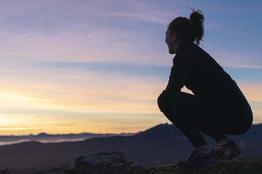 Junge Frau hockt bei Sonnenaufgang auf einem Hügel - SNF01418
