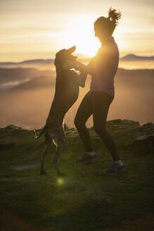 Frau spielt mit Hund auf einem Hügel bei Sonnenaufgang - SNF01432