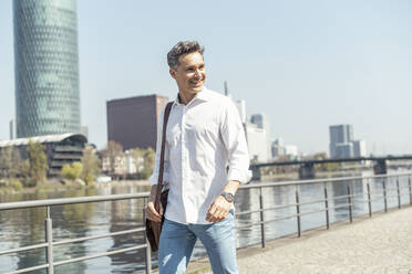 Smiling male entrepreneur with shoulder bag walking on footpath - UUF23158