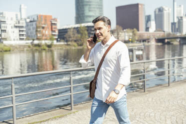 Geschäftsmann mit Umhängetasche, der an einem sonnigen Tag mit seinem Mobiltelefon spricht - UUF23159