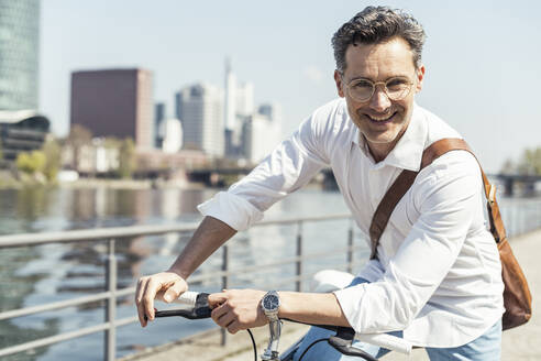 Mature male entrepreneur smiling while leaning on bicycle - UUF23166
