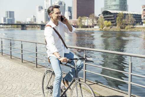 Lächelnder Geschäftsmann auf dem Fahrrad, während er am Fluss in der Stadt mit seinem Handy telefoniert - UUF23170