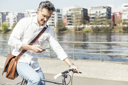 Geschäftsmann, der ein Mobiltelefon benutzt, während er an einem sonnigen Tag Fahrrad fährt - UUF23172