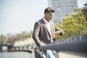 Businessman checking time on wristwatch while leaning on railing - UUF23177