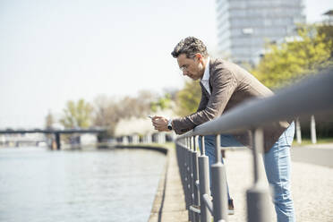 Älterer Geschäftsmann, der ein Mobiltelefon benutzt, während er sich auf ein Geländer am Fluss stützt - UUF23179