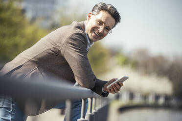 Businessman with mobile phone leaning on railing during sunny day - UUF23180