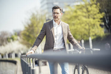 Businessman walking with bicycle by railing on sunny day - UUF23182