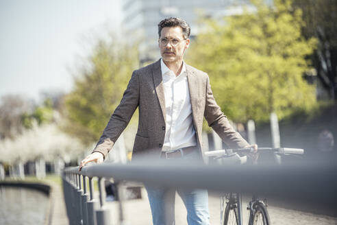 Geschäftsmann zu Fuß mit Fahrrad am Geländer an einem sonnigen Tag - UUF23182