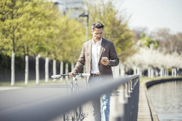 Mature businessman using mobile phone while walking with bicycle by railing - UUF23183