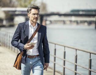 Mature male entrepreneur with crossbody bag and mobile phone looking away while walking by railing - UUF23193